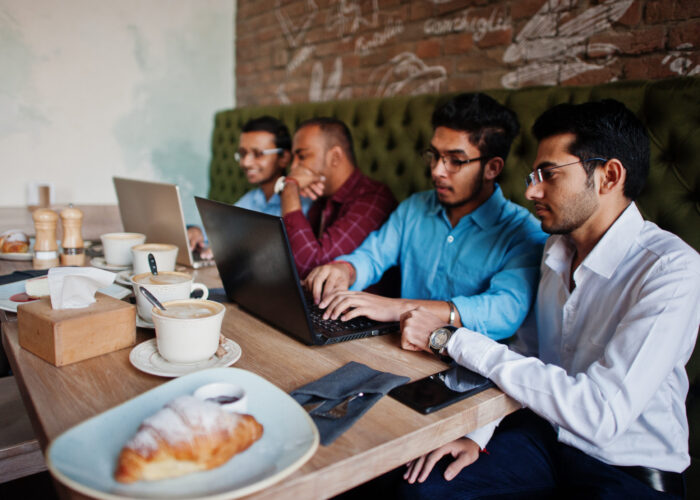 Group of four south asian men's posed at business meeting in cafe. Indians work with laptops together using various gadgets, having conversation.